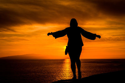 Silhouette woman standing at beach against orange sky