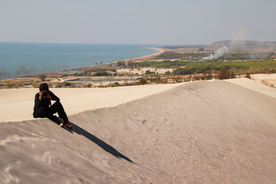 Man photographing while sitting on sand against clear sky