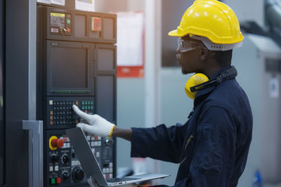 Worker man with yellow helmet and ear protection typing keyboard of laptop computer at factory