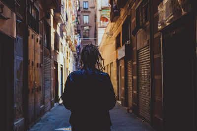 Rear view of man with dreadlocks standing amidst buildings on alley in city