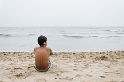 Rear view of shirtless young man sitting at beach against clear sky