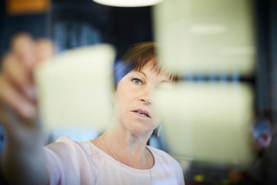 Mature businesswoman sticking adhesive note on glass in creative office