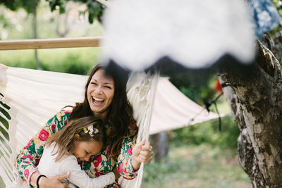 Cheerful mother swinging with daughter on hammock in backyard