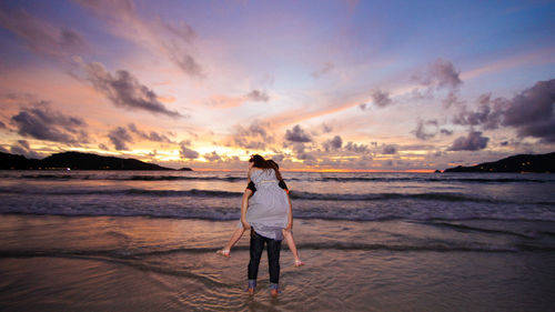 Rear view of boyfriend piggybacking girlfriend at beach against sky during sunset