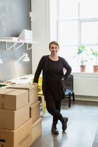 Portrait of smiling businesswoman standing by cardboard boxes stack against window at creative office