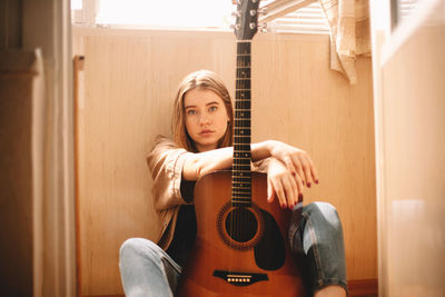 Young woman holding guitar while sitting on balcony floor at home