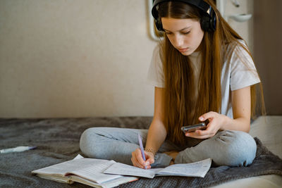Teenage girl at home on bed in headphones and with smartphone, prepares lessons.