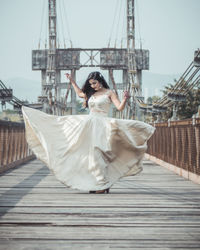 Full length of woman wearing wedding dress standing on footbridge against sky