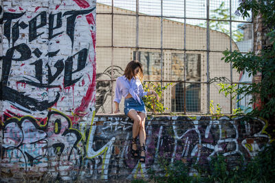 Young woman sitting on window sill amidst graffiti wall