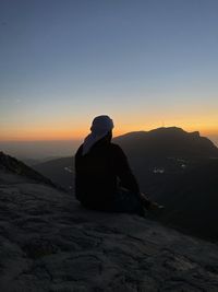 Rear view of silhouette man sitting on rock against sky during sunset