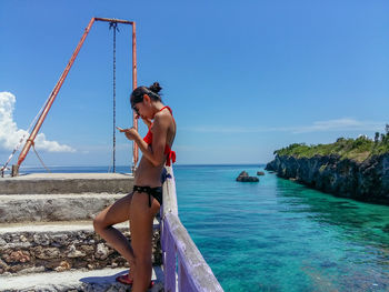 Side view of young woman wearing bikini using phone by sea