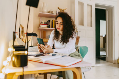 Young curly haired woman using smart phone with books sitting at table