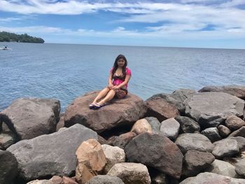 Full length portrait of young woman sitting on rock against sea