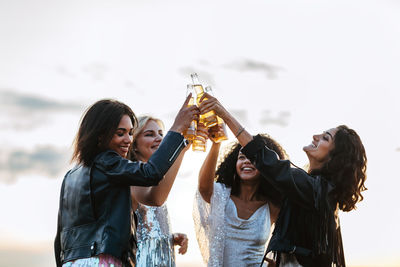 Happy cheerful friends holding beer bottle while standing against sky