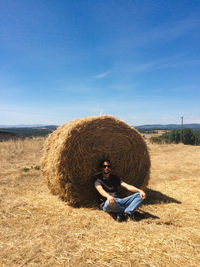 Portrait young man sitting by hay bale against sky