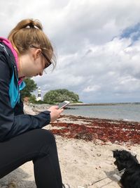 Side view of young woman using mobile phone while sitting by dog at beach against cloudy sky