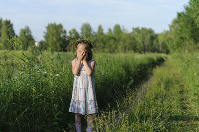 Girl with hands covered face standing on grassy field against sky