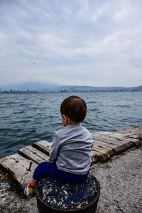 Rear view of boy sitting by sea against sky