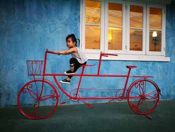 Full length of girl sitting on red artificial bicycle by window