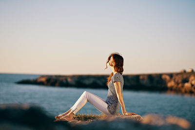 Woman sitting by sea against sky during sunset