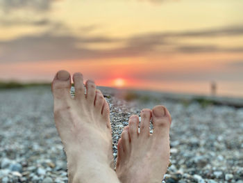 Low section of woman relaxing on beach against sky during sunset