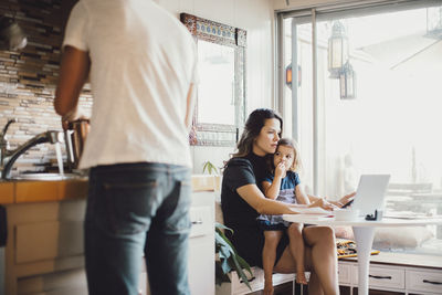 Mother working on laptop while daughter looking at father standing in kitchen