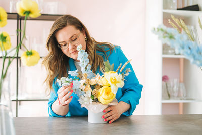 Young woman florist in blue suit and eye glasses with bouquet of flowers in box in flower shop