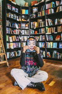 Woman hiding face with book while sitting cross-legged in bookstore