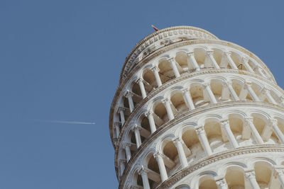 Low angle view of leaning tower against clear blue sky and a jet approaching 