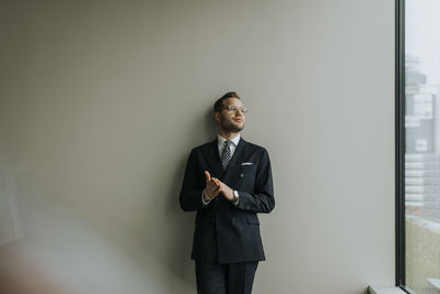 Thoughtful young businessman leaning on gray wall in office