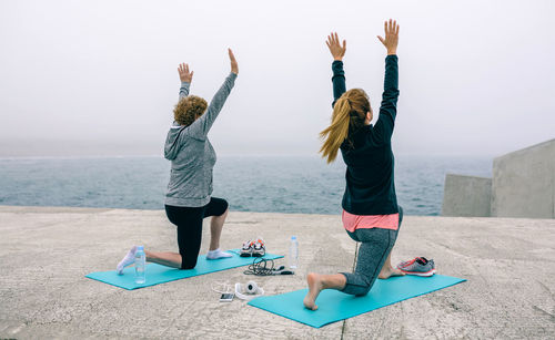 Rear view full length of female friends doing yoga by sea on walkway