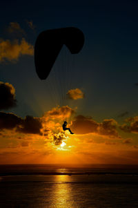 Silhouette person in mid-air paragliding over sea against sky during sunset