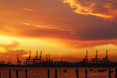 Silhouette cranes by sea against orange sky