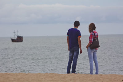 Rear view of friends looking at sailboat in sea while standing at beach