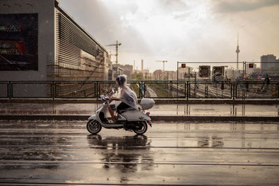 Woman riding motor scooter on road in city against sky