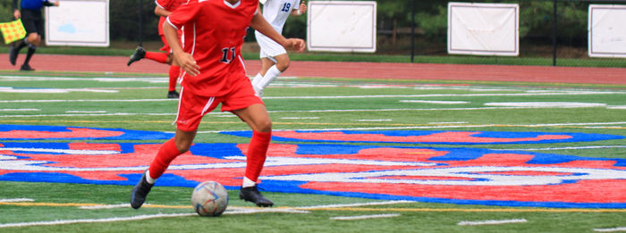 Front view of a high school boy dribbling the soccer ball up field during a game on a turf field.