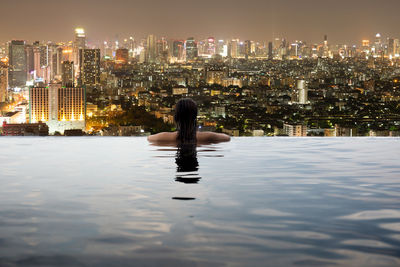 Rear view of woman in swimming pool against illuminated city at night