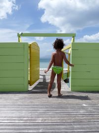 Rear view full length of shirtless child on boardwalk by gate at beach