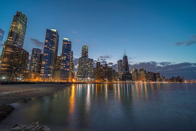 Illuminated buildings by river against sky at dusk