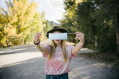 Anonymous female traveler in hat showing disposable medical mask while standing on rural road among forest in autumn day in countryside