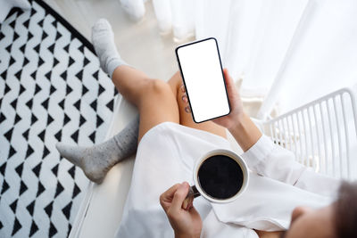 Low section of woman holding coffee cup while using smart phone at home
