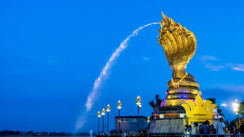 People standing by gold colored fountain against blue sky at dusk