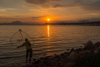 Man fishing in sea against sky during sunset