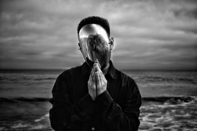 Man in prayer position wearing steel mask at beach against cloudy sky