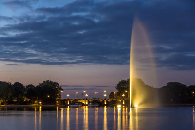 Illuminated building by river against sky at sunset