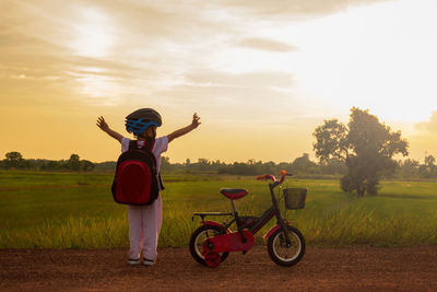 Rear view of boy standing by bicycle on dirt road during sunset