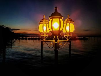 Illuminated lamp by sea against sky at sunset