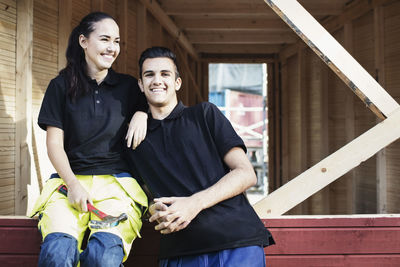 Portrait of happy carpentry student with female friend sitting on wooden cabin