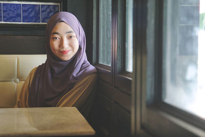 Portrait of young woman wearing hijab while sitting by window at restaurant