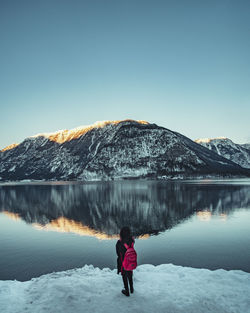 Rear view of woman standing by lake with snowcapped mountain reflection against sky